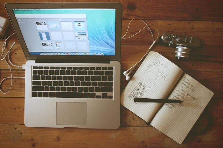 MacBook رمادي متصل بسماعات رأس وشاحن أبيض أمامه كاميرا وكتاب مفتوح بداخله قلم و فيه بعض الكتابات فوق طاولة خشبية 